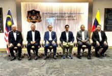 azam - Wilayah.com.my Six men in suits seated in a row for a formal group photo in a government office, with Malaysian and Philippine flags on either side and a crest on the wall behind.