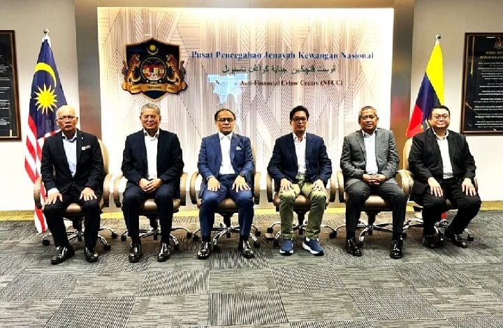 azam - Wilayah.com.my Six men in suits seated in a row for a formal group photo in a government office, with Malaysian and Philippine flags on either side and a crest on the wall behind.