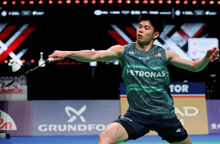 badminton 2 - Wilayah.com.my Male badminton player in a green Petronas jersey stretches to hit a shuttlecock on an indoor court, with sponsor banners in the background.