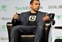 balaji - Wilayah.com.my Man in a navy suit with a black logo T‑shirt, wearing a headset mic, gesturing while seated on stage during a talk.