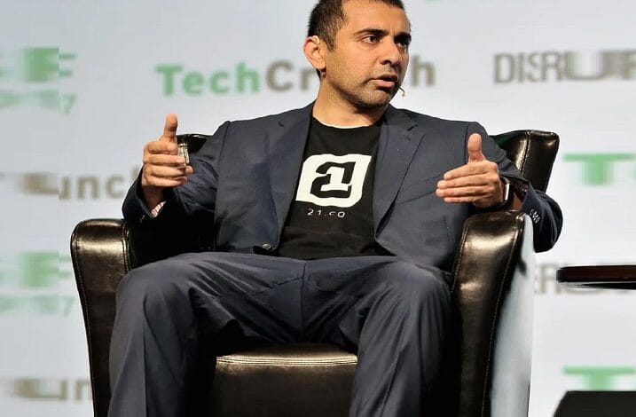 Man in a navy suit with a black logo T‑shirt, wearing a headset mic, gesturing while seated on stage during a talk.