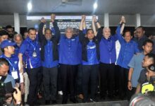 Group of people in blue shirts stand together, raising their arms in a celebratory pose at an indoor event.