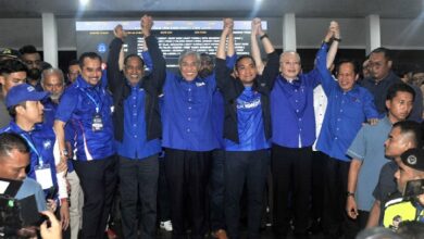 bn - Wilayah.com.my Group of people in blue shirts stand together, raising their arms in a celebratory pose at an indoor event.