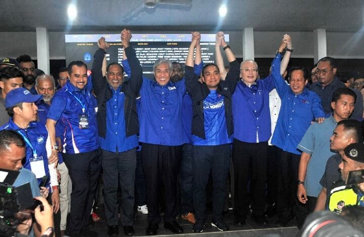 bn - Wilayah.com.my Group of people in blue shirts stand together, raising their arms in a celebratory pose at an indoor event.