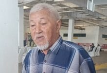 bujang - Wilayah.com.my Elderly man with white hair and goatee, wearing a blue plaid shirt, looking down in a bright indoor hall.