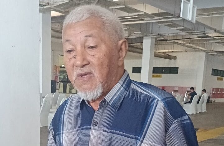 bujang - Wilayah.com.my Elderly man with white hair and goatee, wearing a blue plaid shirt, looking down in a bright indoor hall.