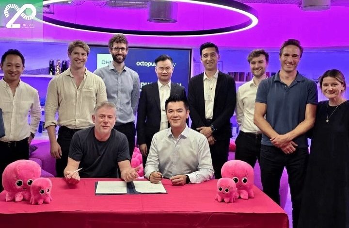 chint - Wilayah.com.my Two men sign an agreement at a pink-clothed table while colleagues stand behind them in a purple-lit office, pink octopus plush toys on the table.