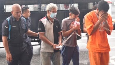 court - Wilayah.com.my Security officers escort two detainees in orange jumpsuits; one detainee covers face as they walk forward with chained hands nearby a masked man and other officer nearby.