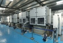 Row of large gray industrial machines connected by hoses in a blue-floored factory hall.