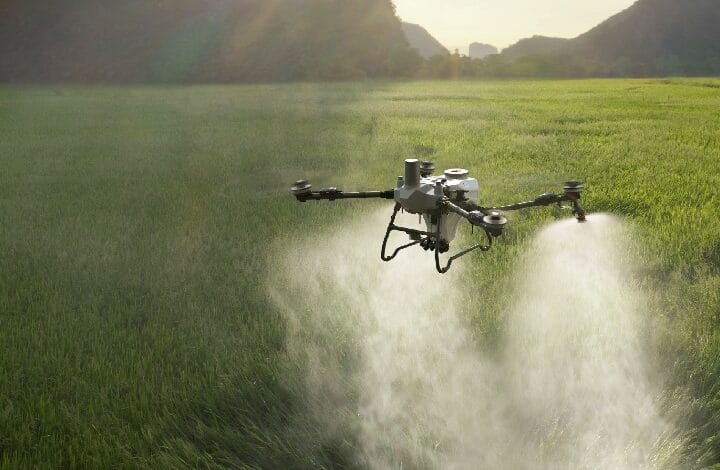 dji 2 - Wilayah.com.my Quadcopter drone spraying a green field, releasing mist over crops at sunset.