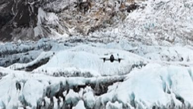 Twin-engine airplane flying low over a pale blue glacier with jagged ice and rocky backdrop.