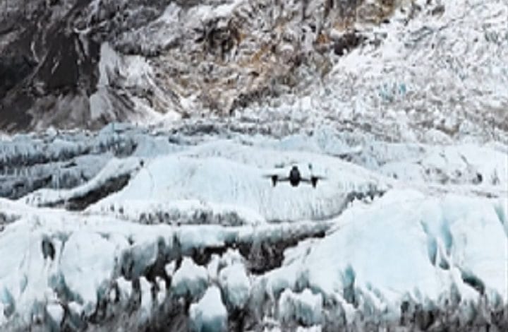 Twin-engine airplane flying low over a pale blue glacier with jagged ice and rocky backdrop.