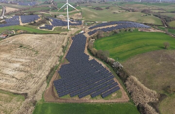 enfinity - Wilayah.com.my Aerial view of a solar farm with rows of panels on rolling hills and a wind turbine in the background.