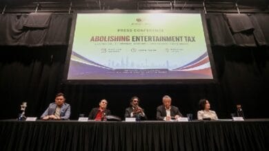 entertaintment act - Wilayah.com.my Panel of five people seated at a long table on a stage during a press conference about abolishing entertainment tax, with a large banner on a screen behind them.