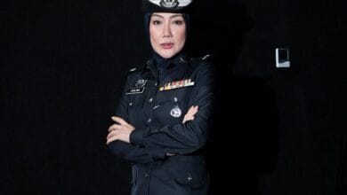 Female police officer in a dark formal uniform with medals, wearing a hijab, arms crossed against a black background.