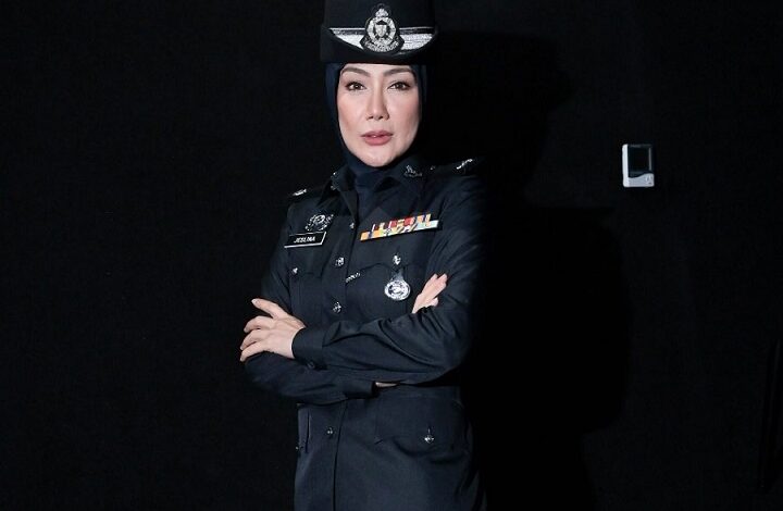 Female police officer in a dark formal uniform with medals, wearing a hijab, arms crossed against a black background.