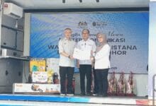 fahmi marina - Wilayah.com.my Three people on a stage during a certificate handover at a formal event; two men in light uniforms present a certificate to a woman wearing a hijab, with a blue banner in the background.