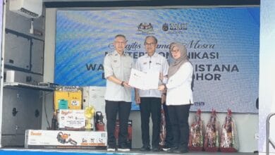 fahmi marina - Wilayah.com.my Three people on a stage during a certificate handover at a formal event; two men in light uniforms present a certificate to a woman wearing a hijab, with a blue banner in the background.