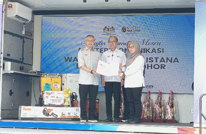 Three people on a stage during a certificate handover at a formal event; two men in light uniforms present a certificate to a woman wearing a hijab, with a blue banner in the background.