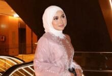 farisha iris 2 - Wilayah.com.my Woman in a pale pink abaya and white hijab posing with a gentle smile in a warmly lit modern interior, decorative embroidery along the chest visible.