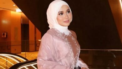 farisha iris 2 - Wilayah.com.my Woman in a pale pink abaya and white hijab posing with a gentle smile in a warmly lit modern interior, decorative embroidery along the chest visible.