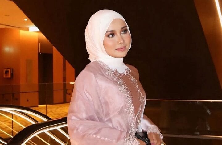 farisha iris 2 - Wilayah.com.my Woman in a pale pink abaya and white hijab posing with a gentle smile in a warmly lit modern interior, decorative embroidery along the chest visible.