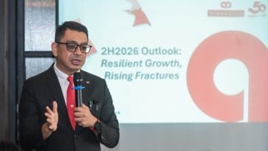 firdaos rosli 2 - Wilayah.com.my Professional man in a dark suit and red tie speaks into a handheld microphone during a presentation, with a slide behind him reading '2H2026 Outlook: Resilient Growth, Rising Fractures'.