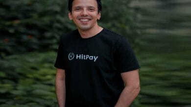 hitpay 2 - Wilayah.com.my Smiling man in a black HitPay T-shirt outdoors with green foliage in the background.