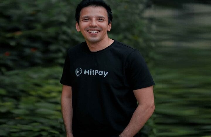 Smiling man in a black HitPay T-shirt outdoors with green foliage in the background.