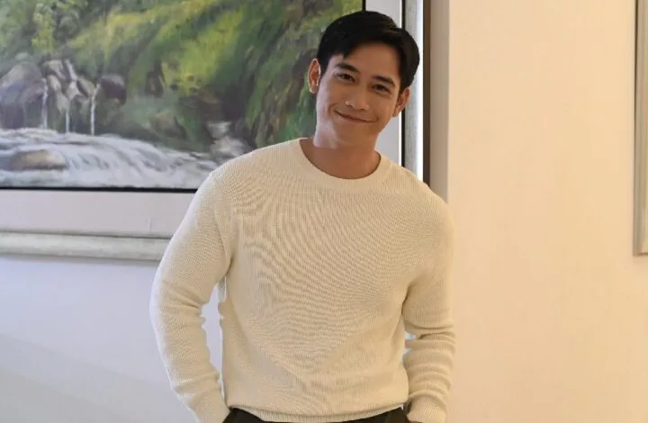 hun haqeem 2 - Wilayah.com.my Man in a white knit sweater smiles at the camera indoors, with a landscape painting on the wall behind him.