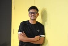 jay iswazir 2 - Wilayah.com.my Young man with glasses and a black T-shirt stands against a bright yellow wall with arms crossed, smiling at the camera.