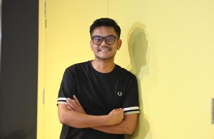 jay iswazir 2 - Wilayah.com.my Young man with glasses and a black T-shirt stands against a bright yellow wall with arms crossed, smiling at the camera.