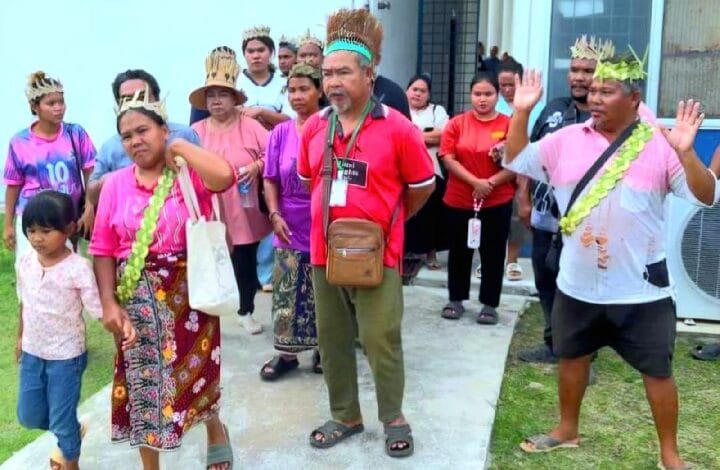 juliana ismail - Wilayah.com.my Group of people in festive attire gathered outside, some wearing feathered headbands; a man in red shirt speaks or leads the activity.