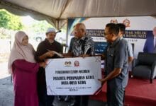 keda - Wilayah.com.my Group of people under a canopy participate in a ceremonial handover, presenting a large sign/banner to a man in a patterned shirt during a community housing grant event in Indonesia.