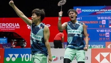 khai xing - Wilayah.com.my Two male badminton players in blue Petronas jerseys celebrate on a court; one raises his racket in triumph as a scoreboard and bright banners loom in the background.