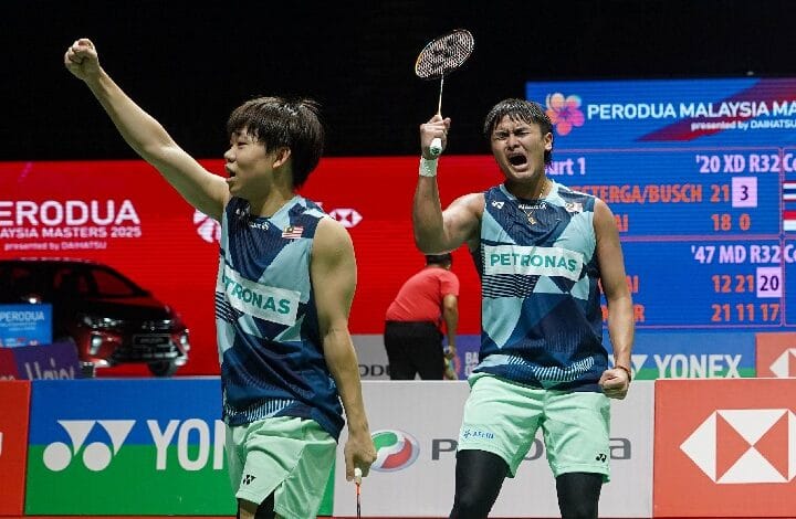 khai xing - Wilayah.com.my Two male badminton players in blue Petronas jerseys celebrate on a court; one raises his racket in triumph as a scoreboard and bright banners loom in the background.