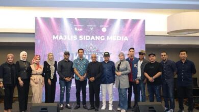 Group of men and women standing in a line on a stage in front of a purple backdrop that reads 'Majlis Sidang Media' during a media briefing.