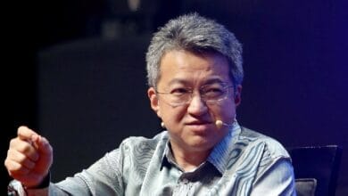 liew chin tong 2 - Wilayah.com.my Man with gray hair wearing a striped blue shirt sits on a stage, speaking into a headset microphone and gesturing with his hand.