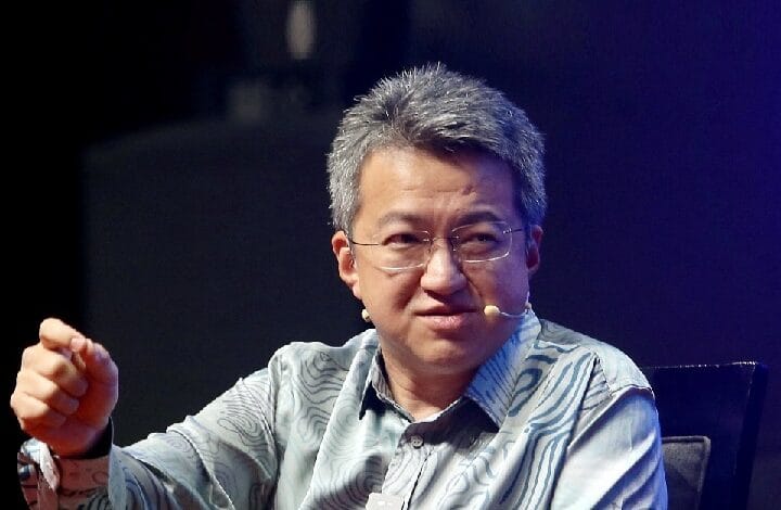 liew chin tong 2 - Wilayah.com.my Man with gray hair wearing a striped blue shirt sits on a stage, speaking into a headset microphone and gesturing with his hand.