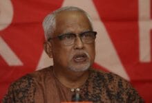 Older man with gray hair and glasses speaking at a podium against a red backdrop with banners behind him.