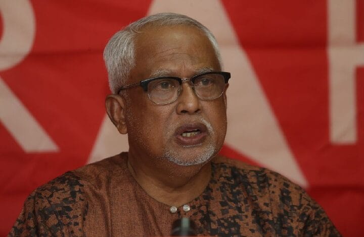 mahfuz - Wilayah.com.my Older man with gray hair and glasses speaking at a podium against a red backdrop with banners behind him.