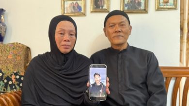 Couple dressed in black sits on a living room couch, holding a smartphone displaying a portrait of a young man, with family photos on the wall behind them.