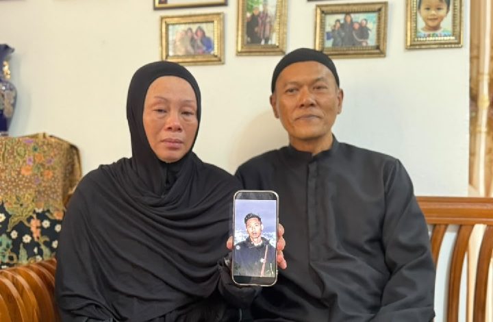 Couple dressed in black sits on a living room couch, holding a smartphone displaying a portrait of a young man, with family photos on the wall behind them.