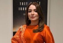 marzella sigai - Wilayah.com.my Portrait of a woman with shoulder-length brown hair, wearing an orange satin top and a red patterned scarf, posing indoors.