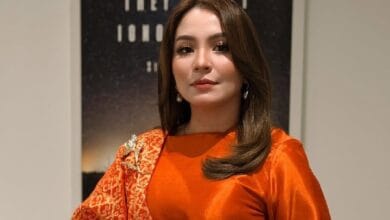 marzella sigai - Wilayah.com.my Portrait of a woman with shoulder-length brown hair, wearing an orange satin top and a red patterned scarf, posing indoors.