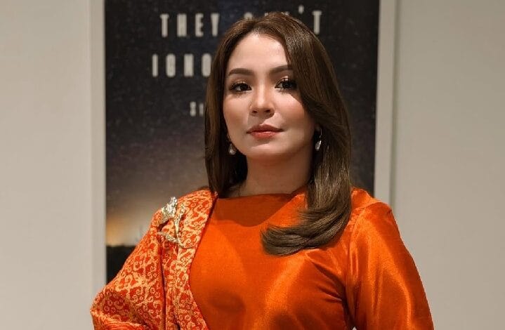 marzella sigai - Wilayah.com.my Portrait of a woman with shoulder-length brown hair, wearing an orange satin top and a red patterned scarf, posing indoors.
