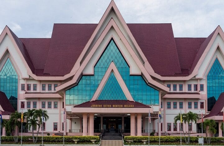 melaka - Wilayah.com.my Front view of a pink government building with steep gabled roofs and blue glass windows; entrance reads 'Jabatan Ketua Menteri Melaka'.