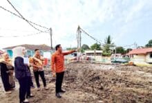 mohd azlan - Wilayah.com.my Man in an orange shirt points toward a cleared construction lot as a small group watches, with houses and utility poles in the background.