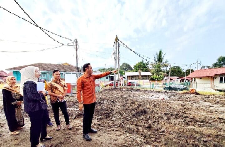 mohd azlan - Wilayah.com.my Man in an orange shirt points toward a cleared construction lot as a small group watches, with houses and utility poles in the background.