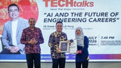 mosti 2 - Wilayah.com.my Three people on a stage during a TEC talks event; a man in patterned shirt receives a book and a bag from a woman in a hijab, with a large screen in the background showing the event title.